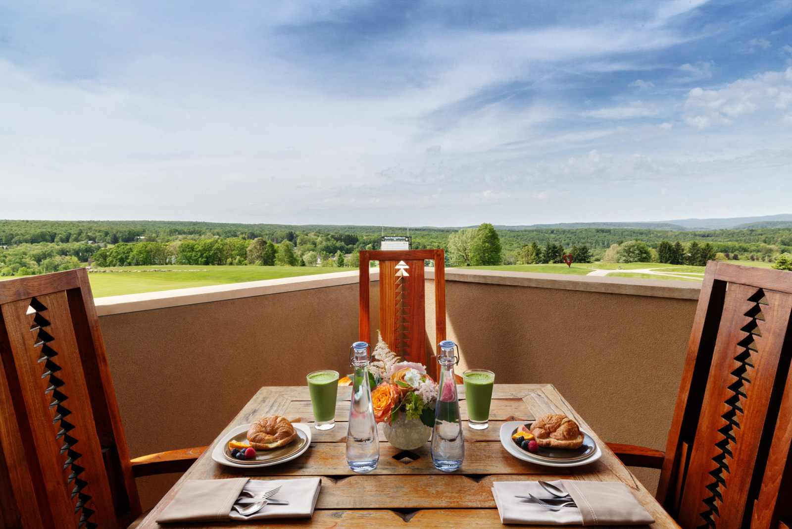 A delightful outdoor dining setup on a private balcony, with wooden chairs and a table set for two, offering a picturesque view of expansive green fields and a bright blue sky.